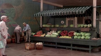 Movie still from “Blaze” (1989), directed by Ron Shelton – A man standing in front of an outdoor fruit stand; Wide shot, High angle