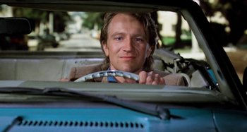 Movie still from “Body Double” (1984), directed by Brian De Palma – A man sitting in the drivers seat of a car holding a steering wheel; Close Up shot, Low angle