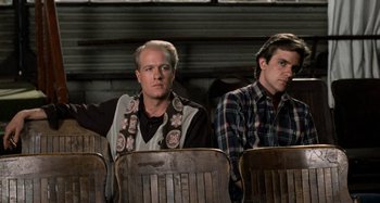 Movie still from “Body Double” (1984), directed by Brian De Palma – Two men sitting next to each other in wooden chairs; Medium shot, High angle