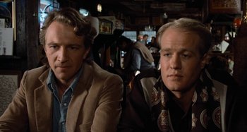 Movie still from “Body Double” (1984), directed by Brian De Palma – Two men sitting at a table in front of a crowd; Close Up shot, Over the shoulder angle