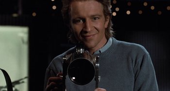Movie still from “Body Double” (1984), directed by Brian De Palma – A man holding a camera in front of a dark background; Close Up shot, Low angle