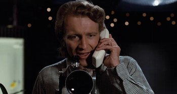Movie still from “Body Double” (1984), directed by Brian De Palma – A man holding a phone up to his ear while standing next to a camera; Close Up shot, Low angle