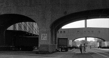 Movie still from “Broadway Danny Rose” (1984), directed by Woody Allen – A man standing under an overpass in a black and white photo; Extreme Wide shot, Low angle