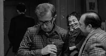 Movie still from “Broadway Danny Rose” (1984), directed by Woody Allen – A man with glasses is holding an object in front of a doll; Medium shot, Over the shoulder angle