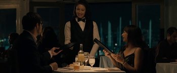 Movie still from “Celeste & Jesse Forever” (2012), directed by Lee Toland Krieger – A woman standing at a dinner table with two people; Medium shot, Over the shoulder angle
