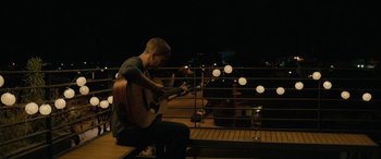 Movie still from “Celeste & Jesse Forever” (2012), directed by Lee Toland Krieger – A man playing a guitar on a balcony at night; Wide shot, Low angle