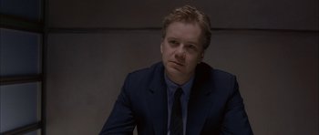Movie still from “Code 46” (2003), directed by Michael Winterbottom – A man wearing a suit and tie sitting in a room; Close Up shot, Over the shoulder angle