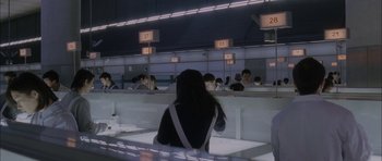 Movie still from “Code 46” (2003), directed by Michael Winterbottom – A woman sitting in front of a group of people in a room; Wide shot, High angle