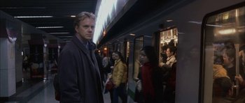 Movie still from “Code 46” (2003), directed by Michael Winterbottom – A man standing in front of a group of people on a train platform; Medium shot, Over the shoulder angle