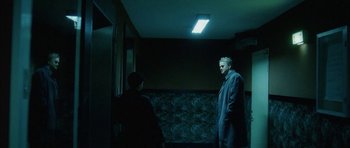 Movie still from “Code 46” (2003), directed by Michael Winterbottom – Two men standing in a dark room , one of whom is wearing a hat; Wide shot, Low angle