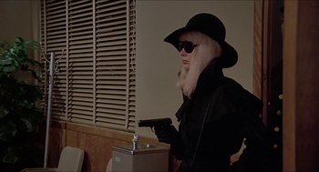 Movie still from “Family Plot” (1976), directed by Alfred Hitchcock – A woman in a black coat and hat holding a gun; Medium shot, Over the shoulder angle