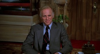 Movie still from “Family Plot” (1976), directed by Alfred Hitchcock – A man in a suit and tie sitting in a chair; Medium shot, Low angle