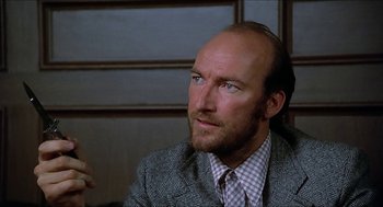 Movie still from “Family Plot” (1976), directed by Alfred Hitchcock – A man in a suit and tie sitting in front of a wall; Close Up shot, Low angle