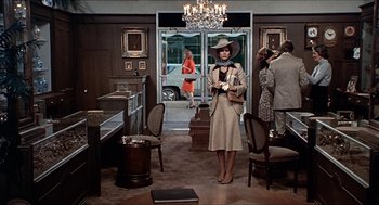 Movie still from “Family Plot” (1976), directed by Alfred Hitchcock – A woman in a hat and a dress standing in front of a door way; Wide shot, Over the shoulder angle