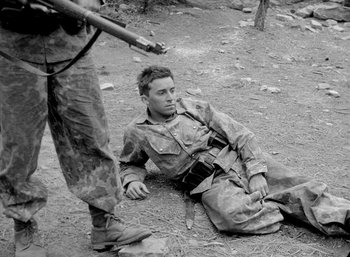 Movie still from “Fear and Desire” (1952), directed by Stanley Kubrick – A man in fatigues sitting on the ground next to another man with a rifle; Medium shot, High angle