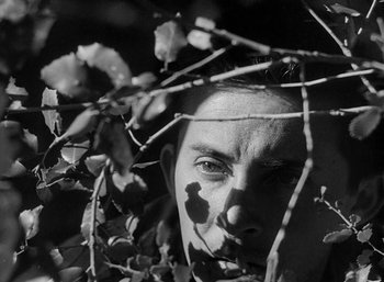 Movie still from “Fear and Desire” (1952), directed by Stanley Kubrick – A black - and - white photo of a man's face behind a tree; Extreme Close Up shot, Low angle