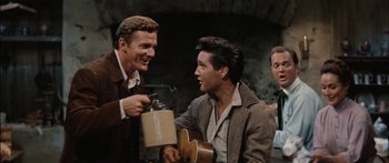 Movie still from “Flaming Star” (1960), directed by Don Siegel – A man holding a guitar while another man holds a jar; Medium shot, Over the shoulder angle