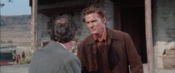 Movie still from “Flaming Star” (1960), directed by Don Siegel – A man wearing a brown jacket talking to another man in front of a brick wall; Medium shot, Over the shoulder angle