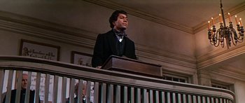 Movie still from “Hawaii” (1966), directed by George Roy Hill – A man in a suit and tie standing at a podium in front of an audience; Medium shot, Low angle
