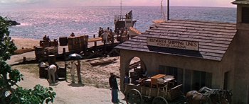 Movie still from “Hawaii” (1966), directed by George Roy Hill – People are standing on the beach near a pier; Extreme Wide shot, High angle