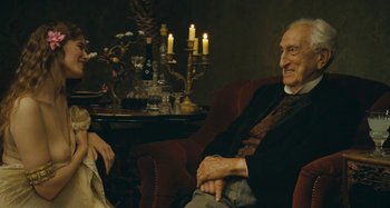 Movie still from “House of Tolerance” (2011), directed by Bertrand Bonello – An older man and a woman sitting in a chair; Medium shot, Over the shoulder angle