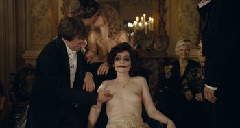 Movie still from “House of Tolerance” (2011), directed by Bertrand Bonello – A woman is sitting in a chair with a man and two other women; Medium shot, Over the shoulder angle
