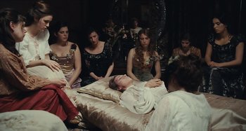 Movie still from “House of Tolerance” (2011), directed by Bertrand Bonello – A group of people sitting around a bed with a man laying on it; Medium shot, High angle