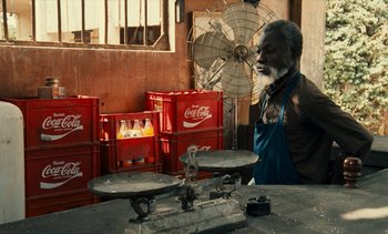 Movie still from “Hyenas” (1992), directed by Djibril Diop Mambéty – An old man standing in front of a table; Medium shot, High angle
