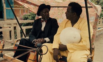 Movie still from “Hyenas” (1992), directed by Djibril Diop Mambéty – Two men sitting next to each other on a bench; Medium shot, Over the shoulder angle