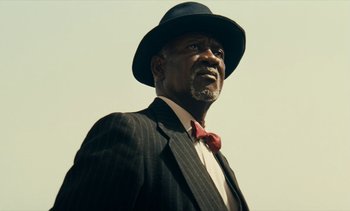 Movie still from “Hyenas” (1992), directed by Djibril Diop Mambéty – An older man in a suit and a red tie; Close Up shot, Low angle