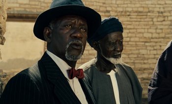 Movie still from “Hyenas” (1992), directed by Djibril Diop Mambéty – Two older black men standing next to each other in front of a brick wall; Close Up shot, Low angle
