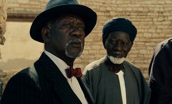 Movie still from “Hyenas” (1992), directed by Djibril Diop Mambéty – Two older men standing next to each other in front of a brick wall; Close Up shot, Low angle
