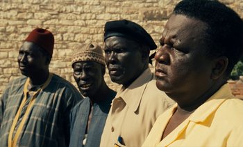 Movie still from “Hyenas” (1992), directed by Djibril Diop Mambéty – A group of men standing next to each other in front of a brick wall; Close Up shot, High angle