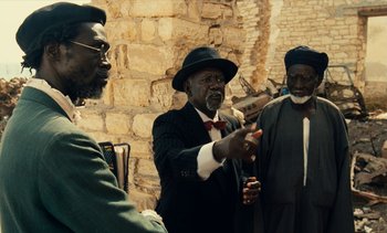 Movie still from “Hyenas” (1992), directed by Djibril Diop Mambéty – A group of men standing next to each other in front of a brick wall; Medium shot, Over the shoulder angle