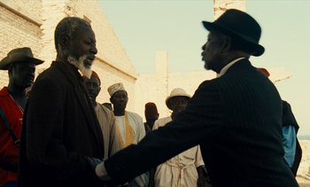Movie still from “Hyenas” (1992), directed by Djibril Diop Mambéty – Two men shaking hands in front of a group of people; Medium shot, Low angle
