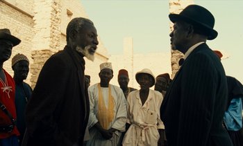 Movie still from “Hyenas” (1992), directed by Djibril Diop Mambéty – A group of men standing next to each other in front of a brick wall; Medium shot, Over the shoulder angle