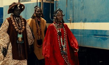 Movie still from “Hyenas” (1992), directed by Djibril Diop Mambéty – A woman in a red dress and a man in a black suit; Medium shot, Low angle