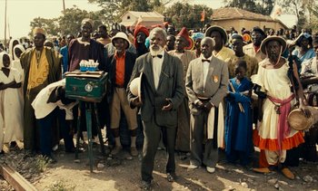 Movie still from “Hyenas” (1992), directed by Djibril Diop Mambéty – An old man standing in front of a group of people; Wide shot, High angle