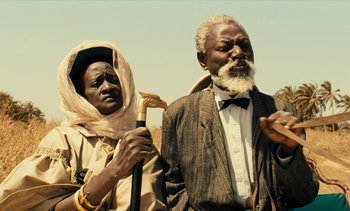 Movie still from “Hyenas” (1992), directed by Djibril Diop Mambéty – A man and a woman standing next to each other; Medium shot, Low angle