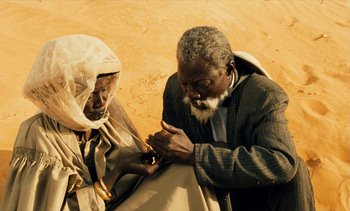 Movie still from “Hyenas” (1992), directed by Djibril Diop Mambéty – An older man and woman are sitting on the ground; Medium shot, High angle