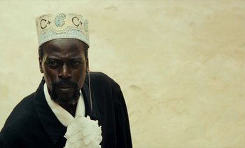 Movie still from “Hyenas” (1992), directed by Djibril Diop Mambéty – A man wearing a black robe and a crown; Close Up shot, Low angle