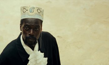 Movie still from “Hyenas” (1992), directed by Djibril Diop Mambéty – A man wearing a crown and holding his hands together; Close Up shot, Low angle