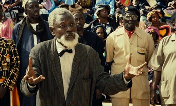 Movie still from “Hyenas” (1992), directed by Djibril Diop Mambéty – An older man in a suit and a bow tie is standing in front of a group of people; Medium shot, Over the shoulder angle