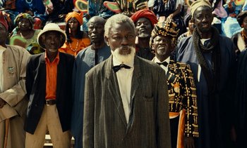 Movie still from “Hyenas” (1992), directed by Djibril Diop Mambéty – An old man standing in front of a group of people; Medium shot, High angle