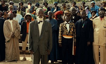 Movie still from “Hyenas” (1992), directed by Djibril Diop Mambéty – A group of men standing in front of each other; Wide shot, Over the shoulder angle