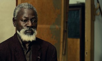 Movie still from “Hyenas” (1992), directed by Djibril Diop Mambéty – An older man with a beard is looking at the camera; Close Up shot, Over the shoulder angle