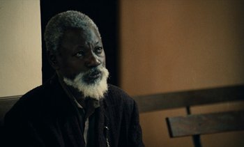 Movie still from “Hyenas” (1992), directed by Djibril Diop Mambéty – An old man with a beard and a white mustache; Close Up shot, Over the shoulder angle
