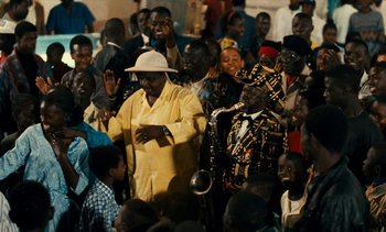 Movie still from “Hyenas” (1992), directed by Djibril Diop Mambéty – A group of people standing around each other in front of a microphone; Medium shot, Over the shoulder angle