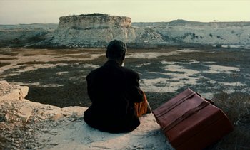Movie still from “Hyenas” (1992), directed by Djibril Diop Mambéty – A person sitting on a rock with a suitcase; Wide shot, High angle
