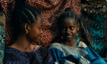 Movie still from “Hyenas” (1992), directed by Djibril Diop Mambéty – Two young women sitting next to each other on a blanket; Medium shot, High angle